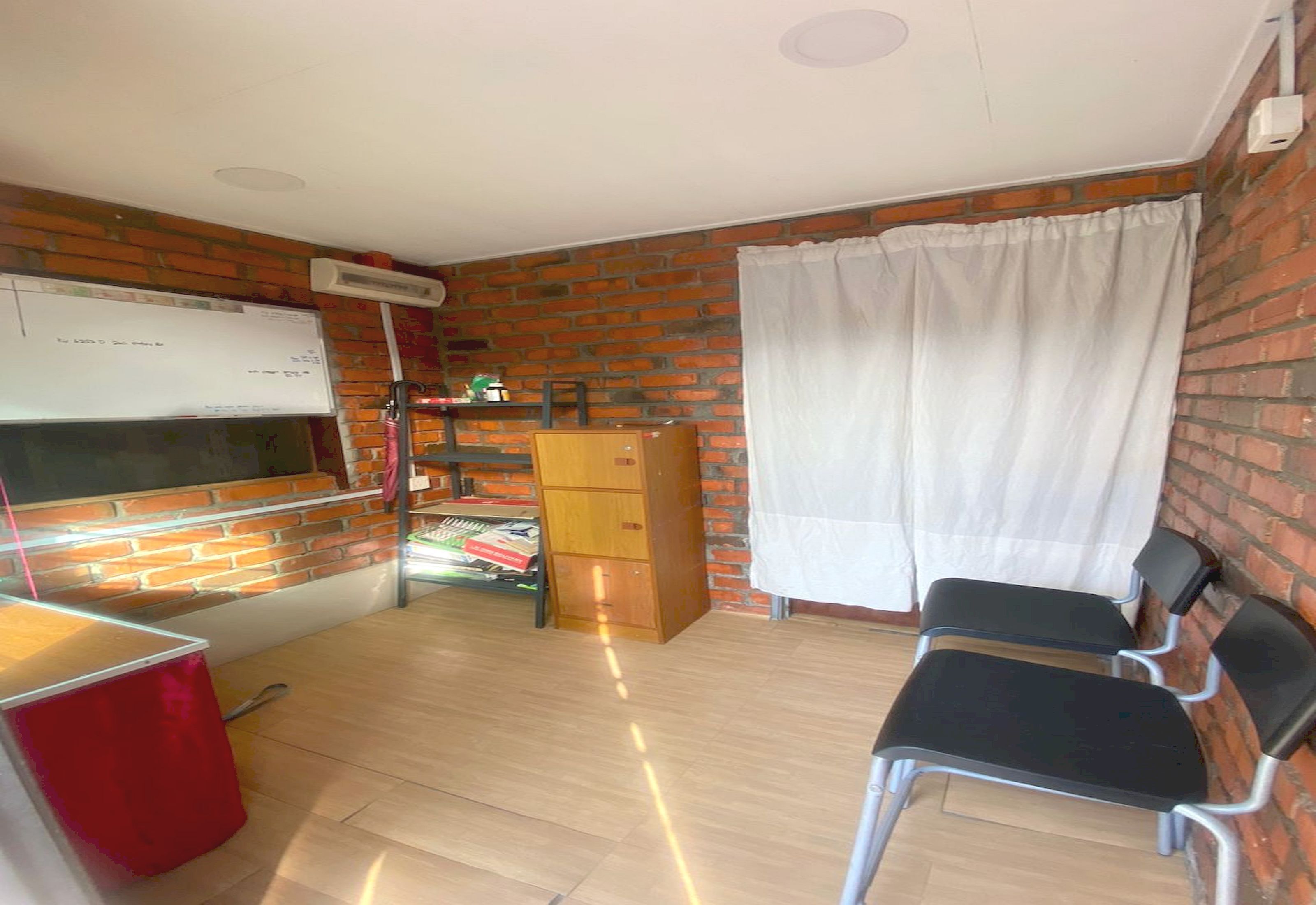 Una habitación con pared de ladrillo, una pizarra, un armario de madera, un bote de basura rojo y dos sillas negras.