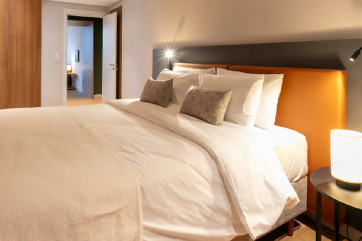 Una cama bien hecha con cabecera, una lámpara de noche y vista a un pasillo.