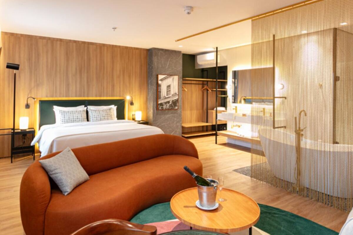 Una habitación de hotel moderna con una cama grande, un sofá curvo, una mesita redonda con una botella y un vaso, y un vestidor con ducha.