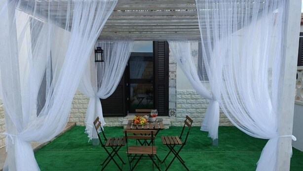 An outdoor seating area with a table and chairs, draped with white fabric, and a stone wall in the background.