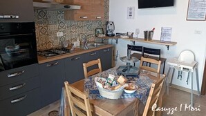 A compact kitchen with a gas stove, oven, and a sink. There is a dining table with chairs and a baby high chair. A television is mounted on the wall, and a shelf with various items is visible.