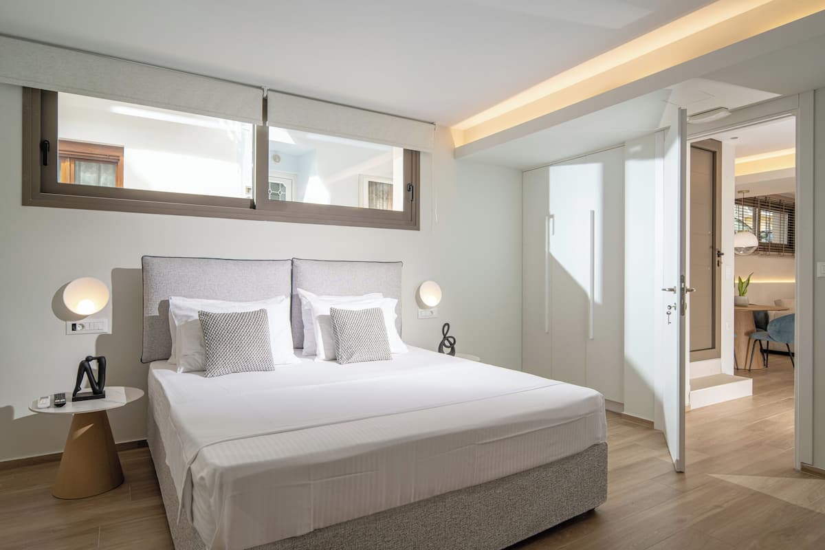 A modern hotel room with a large bed, bedside tables, a mirror, and a view into the adjacent dining area.
