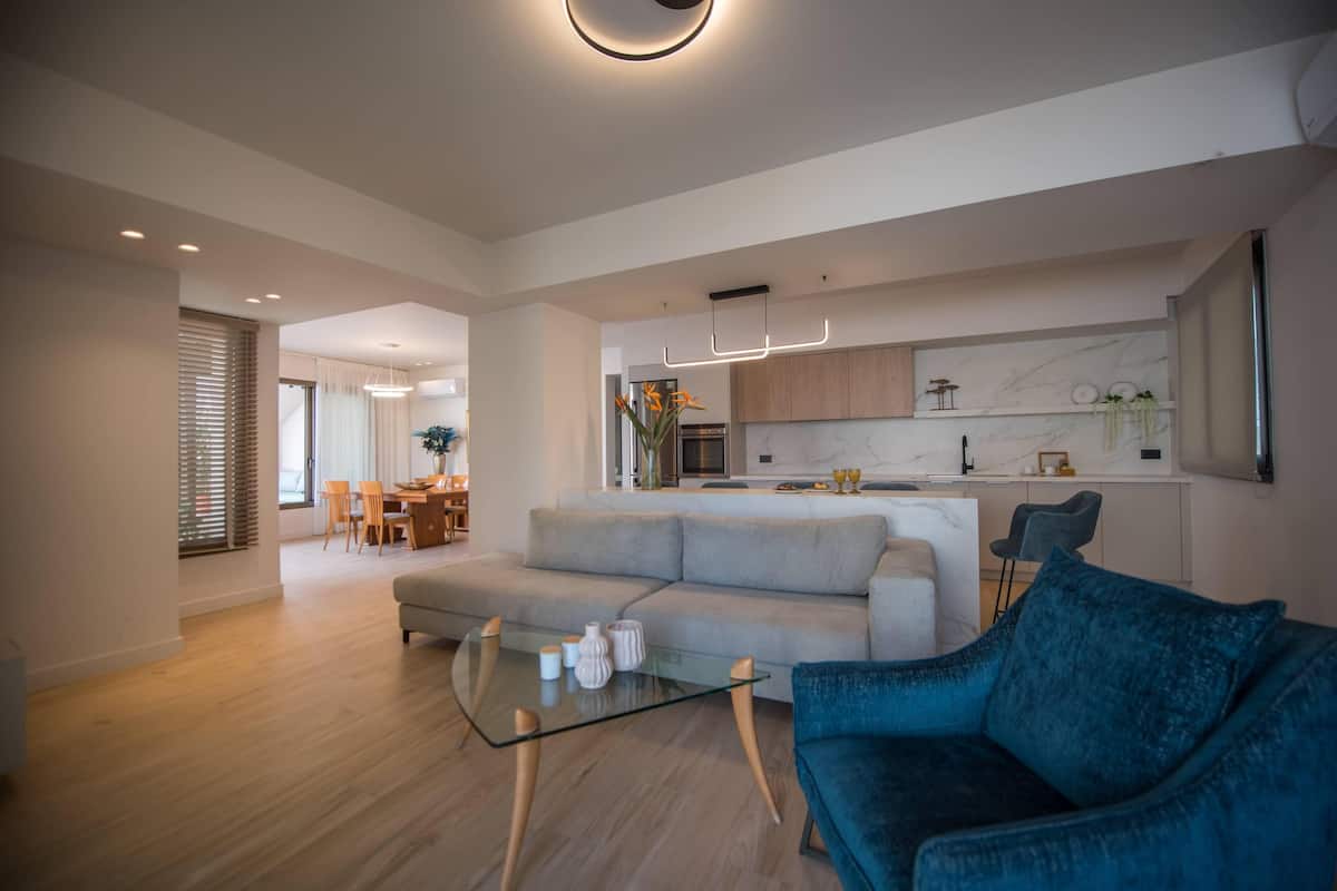 A modern living room with a glass coffee table, a blue sofa, and a kitchen in the background.