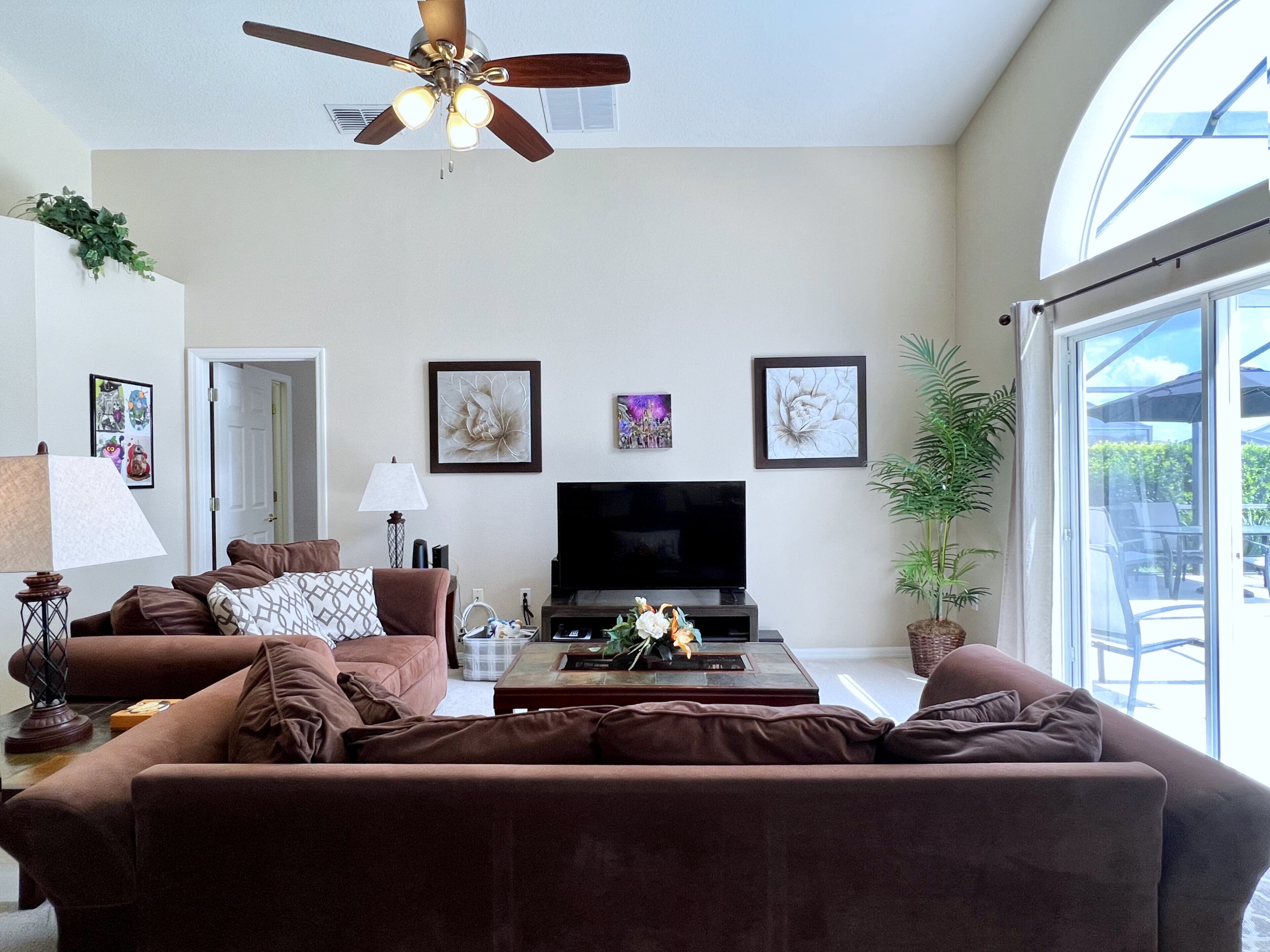 A living room with a ceiling fan, a flat-screen TV, brown sofas, a coffee table, and a large window with a view.