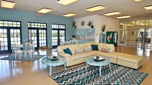 A spacious lobby with a beige sectional sofa, two glass coffee tables, and a patterned rug. There are two white dining tables with chairs, a kitchen area with teal cabinets, and large windows allowing natural light.