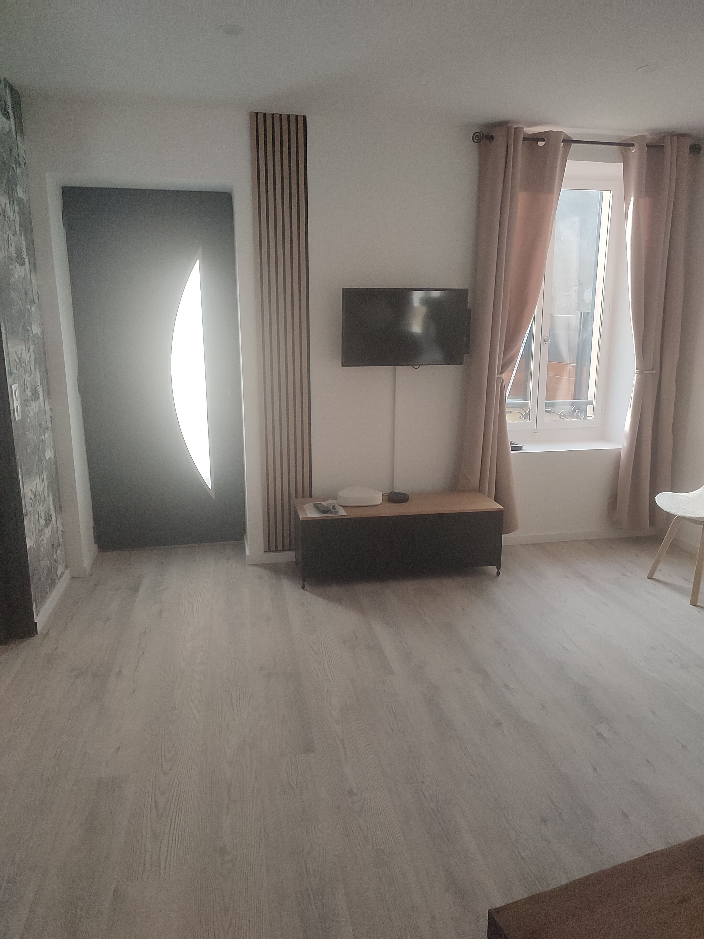 A room with a television on a stand, a bench, a window with curtains, and a door with a light fixture.