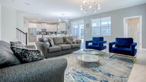 Ein modernes Wohnzimmer mit einem grauen Sofa, blauen Sesseln und einem Glastisch. Im Hintergrund ist eine Küche mit weißen Schränken und einem Edelstahlkühlschrank erkennbar.