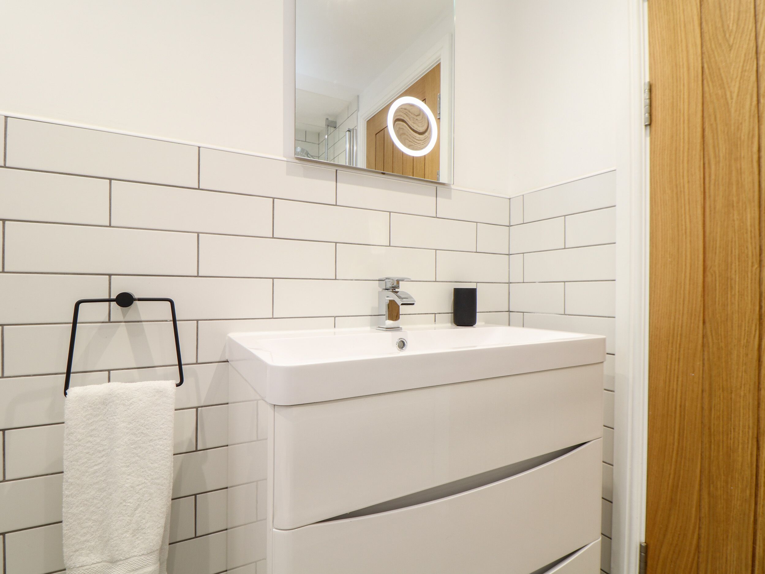 Un bagno moderno rivestito di piastrelle bianche, con un lavandino bianco, uno specchio con illuminazione e una porta in legno.