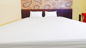 Un lit avec du linge de lit blanc et deux oreillers, une tête de lit en bois ornée d’un motif cachemire et une chaise rouge sur la droite.