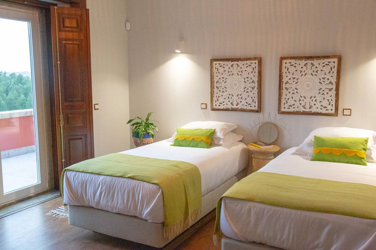 A hotel room with two beds, a potted plant, decorative wall panels, and a view of greenery outside.