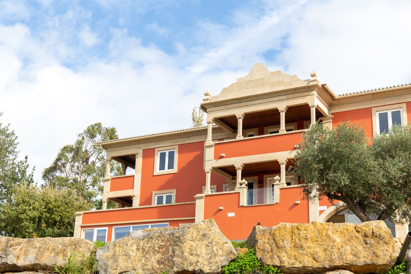 A multi-story building with balconies, large rocks in the foreground, and trees in the background.