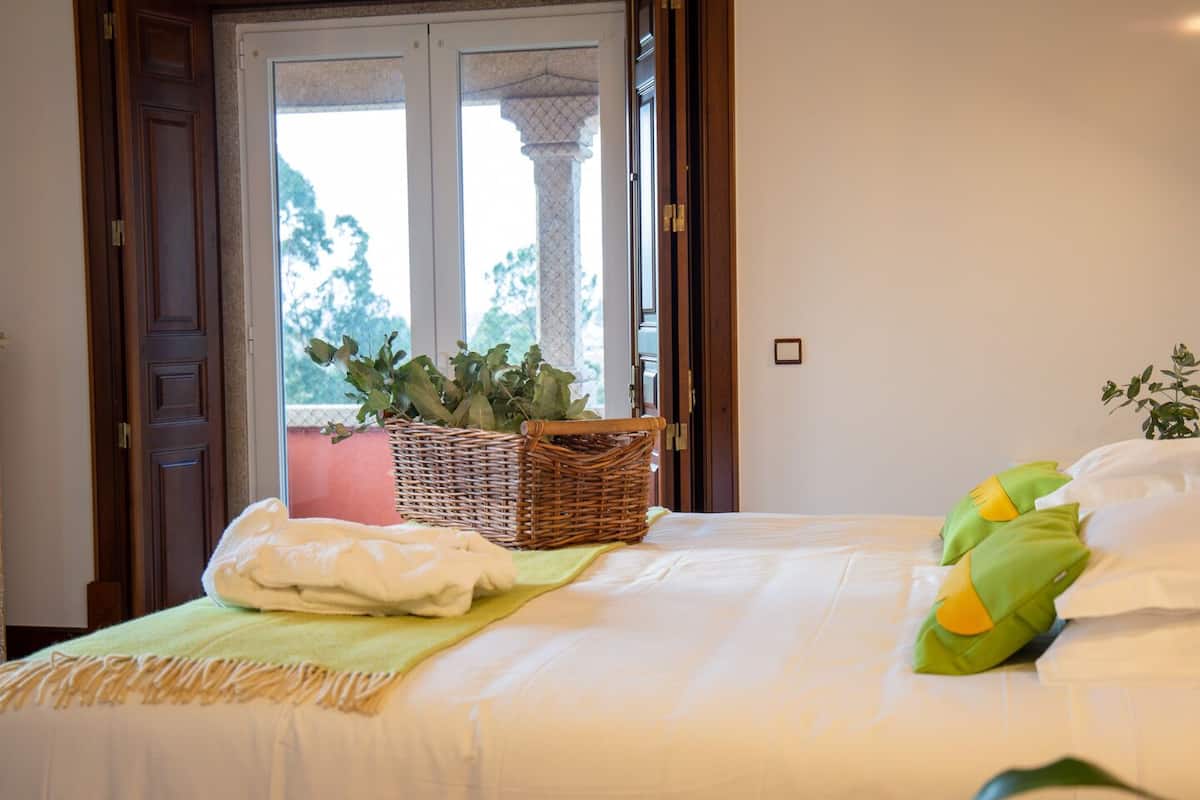 A bedroom with a bed, a wicker basket with plants, and a view of trees through a window.