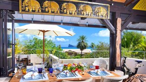 Un área de comedor al aire libre con una mesa de madera dispuesta para cuatro personas, rodeada de sillas de mimbre, un paraguas grande y vistas al océano y palmeras.
