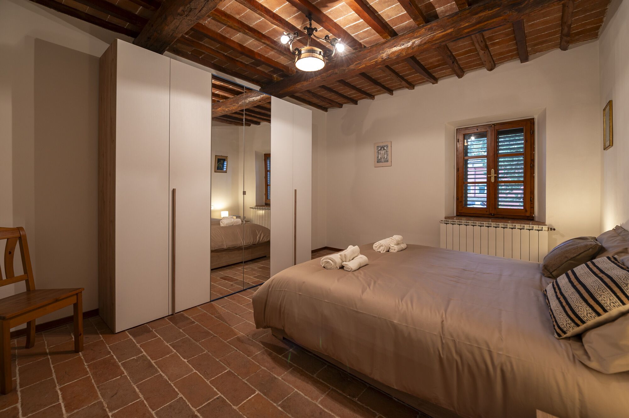 Een slaapkamer met een bed, een houten stoel, een kledingkast, een raam met zonwering en een radiator.