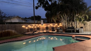 Una piscina con cubierta de madera, sillas de descanso y luces colgantes.