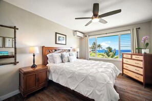 Quarto com cama, mesas de cabeceira, cômoda, ventilador de teto e janela com vista para palmeiras e o oceano.
