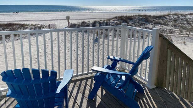 Een houten terras met blauwe Adirondack-stoelen met uitzicht op een strand.