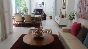 Ein modernes Wohnzimmer mit einem runden Holztisch, einer beigen Couch und einem Essbereich mit Korbstühlen und einem weißen Tisch.