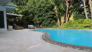 Ein Pool mit gewölbter Kante, umgeben von einem Kiesstreifen. Liegestühle und eine überdachte Sitzgelegenheit befinden sich in der Nähe.