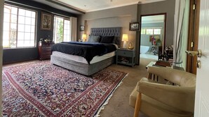 Un dormitorio con una cama grande, una mesita de noche, una silla, una alfombra y una ventana con cortinas.