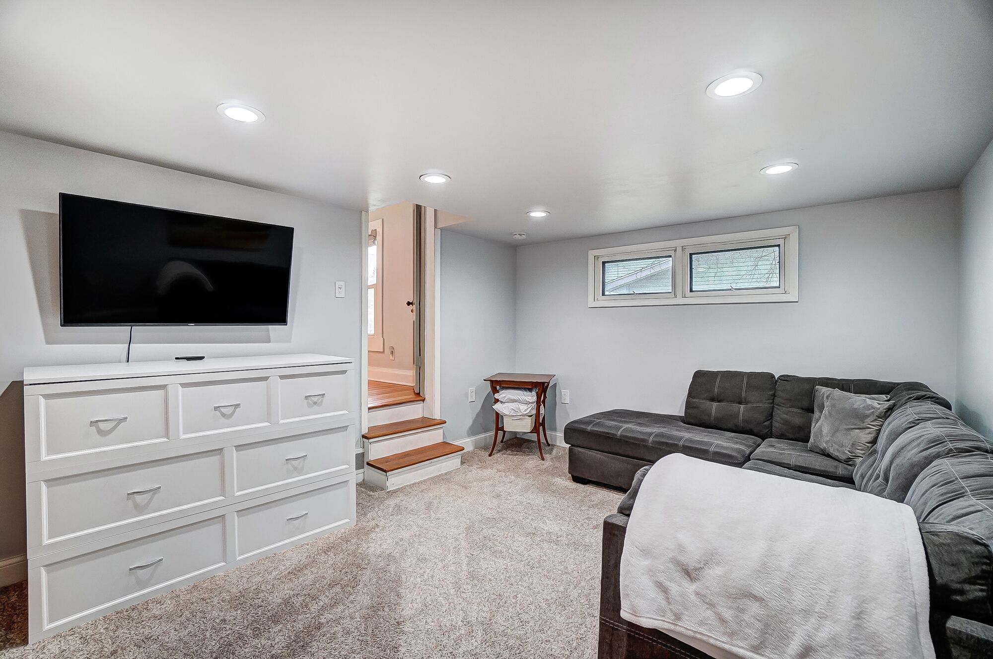 A living room with a TV, white dresser, grey sofa, and a window with a view.