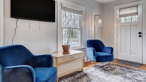 Una sala de estar moderna con un televisor de pantalla plana, dos sillones azules, un aparador de madera y un amplio ventanal con vistas a árboles.