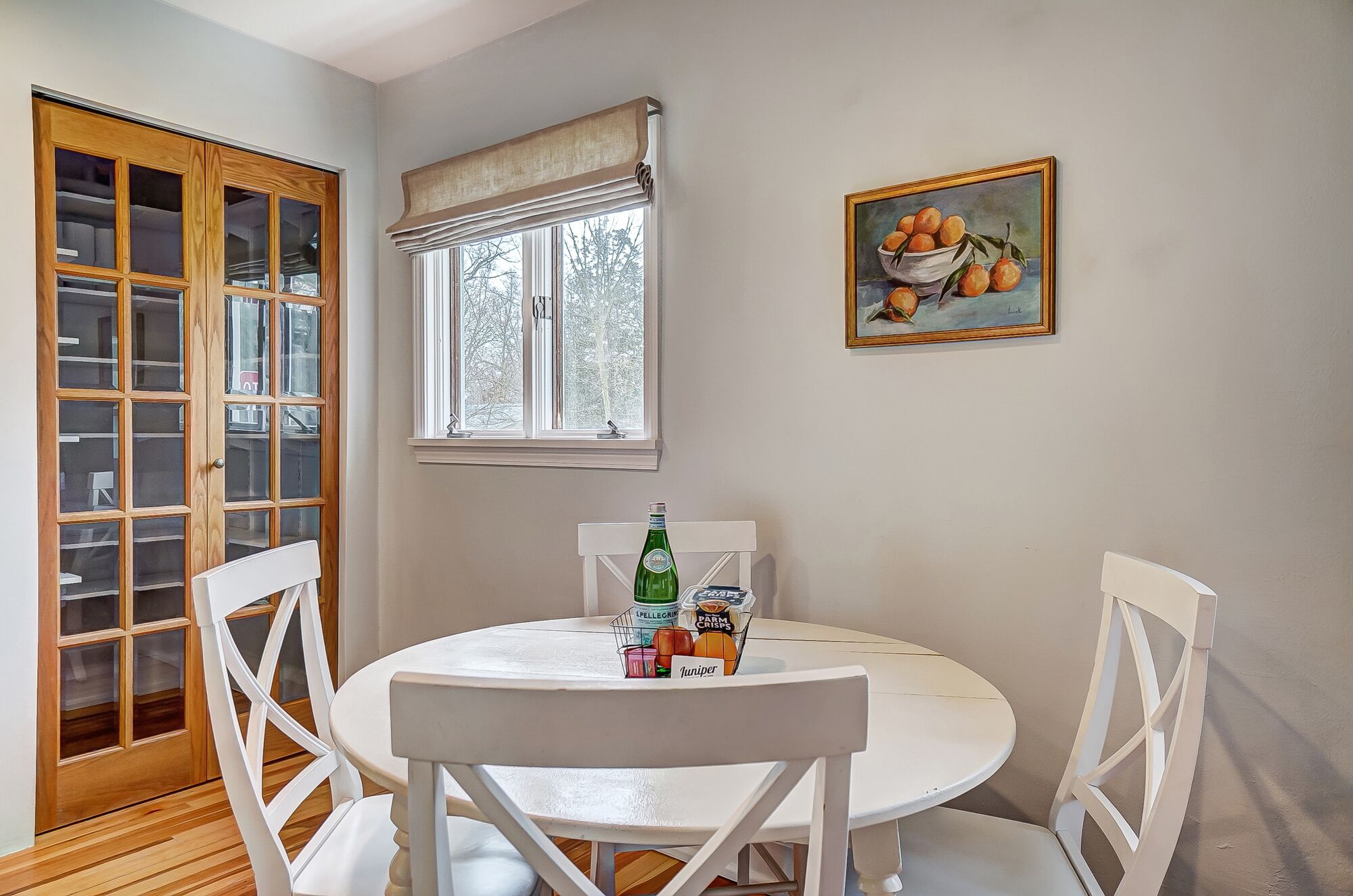 Un comedor con una mesa redonda, sillas blancas, una ventana con cortina, una puerta de madera con paneles de vidrio y un cuadro en la pared.