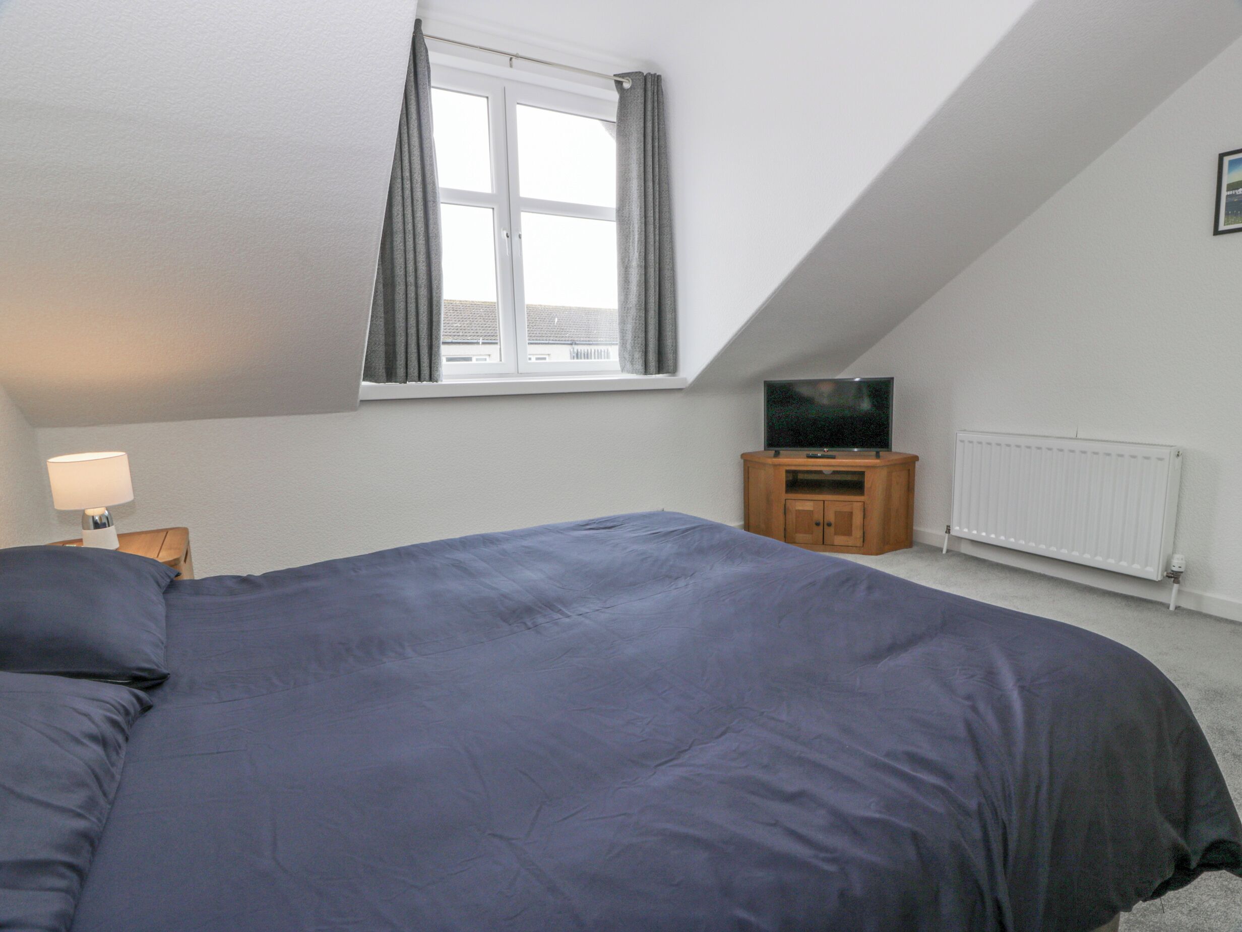A bedroom with a large bed, a TV on a wooden stand, a window with curtains, and a radiator.
