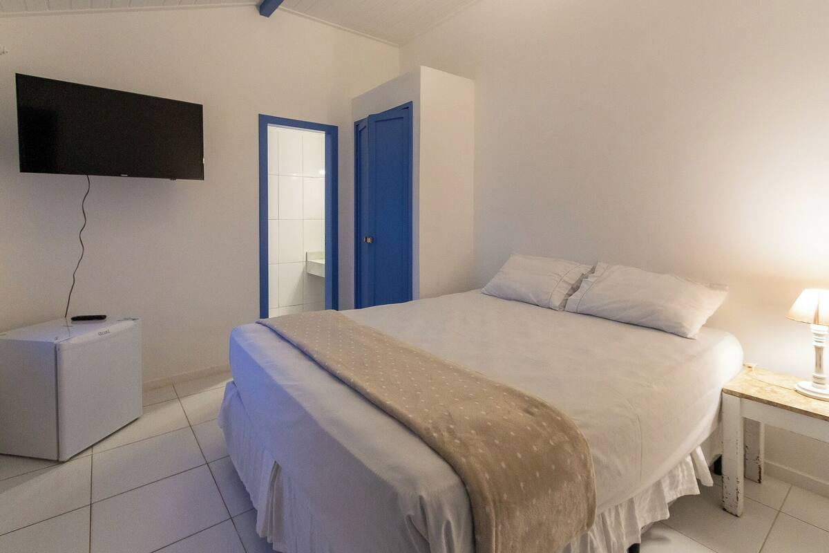 A bedroom with a bed, a TV mounted on the wall, a small bedside table with a lamp, and a door with a blue frame.
