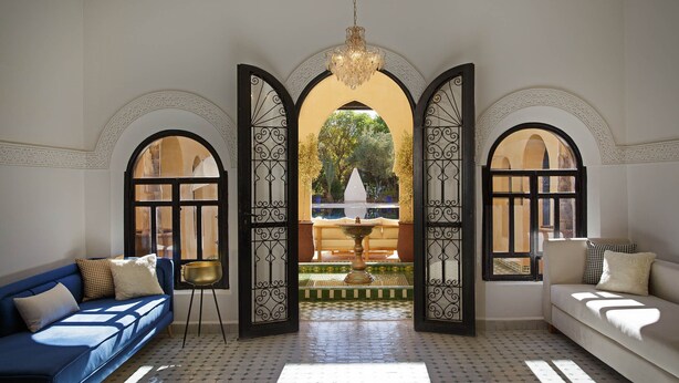 Une pièce avec un canapé bleu, un canapé blanc et un lampadaire doré. Trois portes en arc donnent sur un espace extérieur agrémenté d’une fontaine et d’arbres.