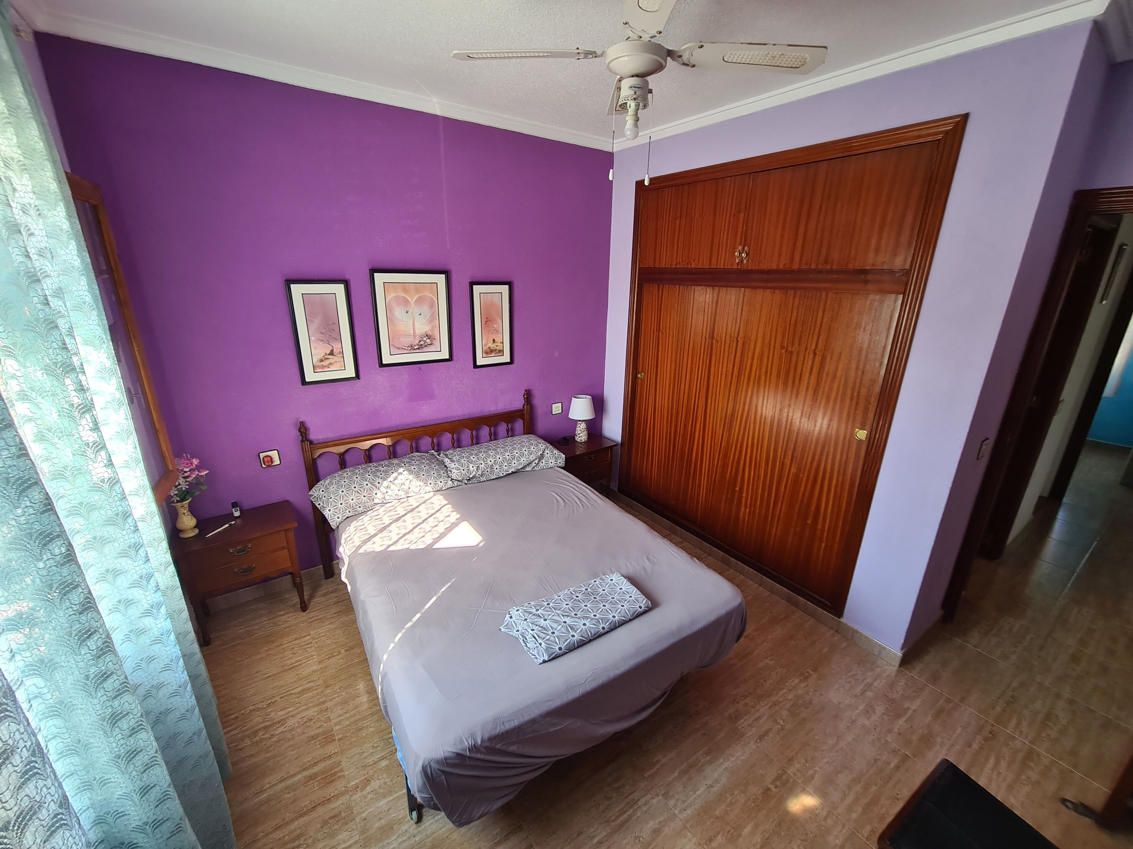 Een slaapkamer met een bed, een houten garderobe, een nachtkastje en een plafondventilator.