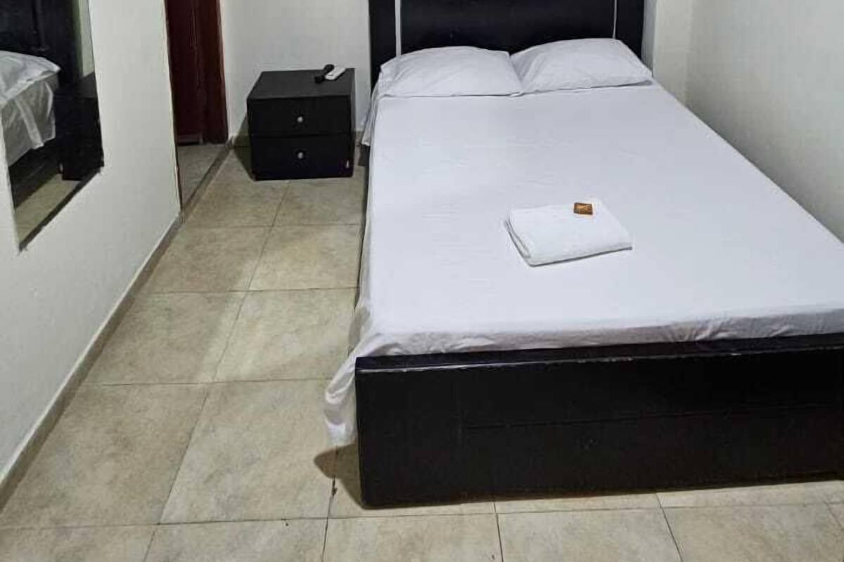 A bedroom with a bed, an air conditioning unit, a nightstand, a suitcase, and a painting on the wall.