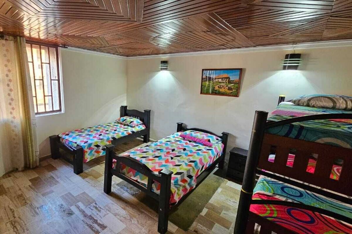 A room with two single beds, a wooden ceiling, a window with curtains, a painting on the wall, and a desk with a chair.