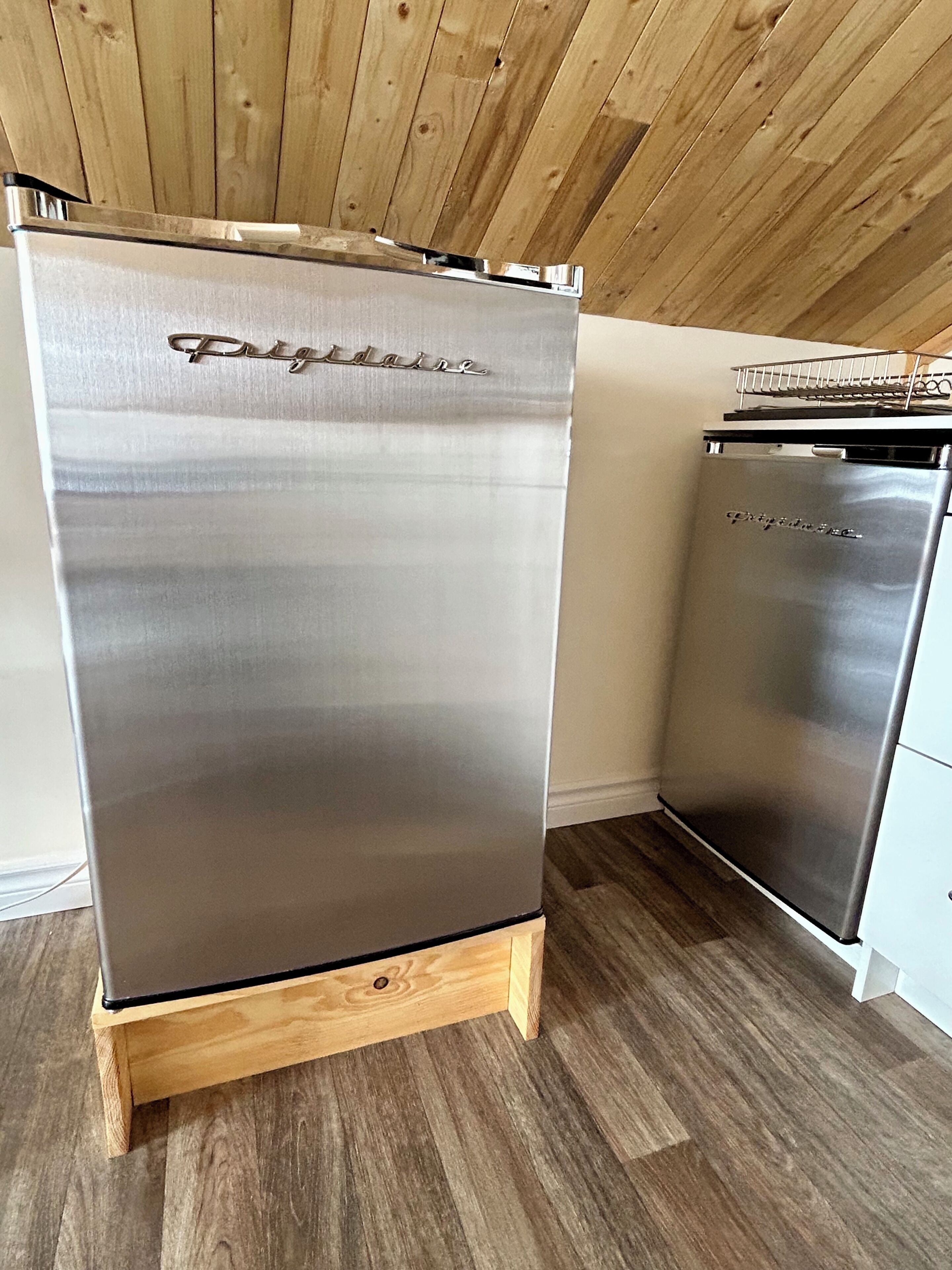 Un réfrigérateur en acier inoxydable, posé sur un socle en bois, dans une cuisine dont le plafond et le sol sont également en bois.