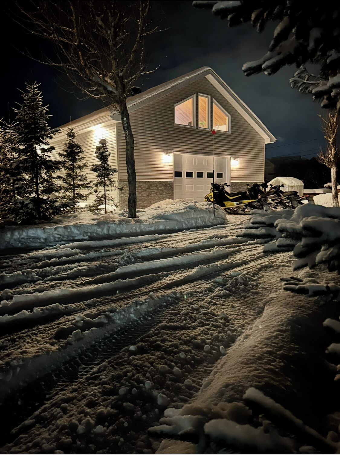 A house with a garage, surrounded by snow-covered ground and trees.
