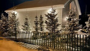 Paesaggio innevato con un edificio bianco, una recinzione di legno e diversi alberi sempreverdi.