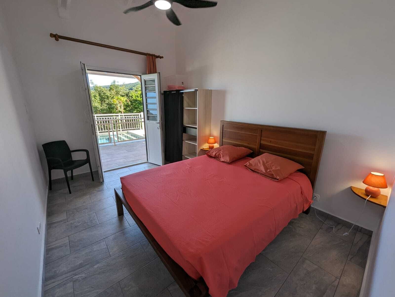 Un dormitorio con cama, una silla, armario, ventilador de techo y balcón con vista a la vegetación.