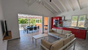 A spacious living area with a sofa, a dining table, and a kitchen with red cabinets. - Villa Tiaré Tahiti (Deshaies)