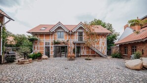 Een tweelaags huis met een pannendak, een met stenen bestraat binnenterrein en een kleine tuin met een boom en een bankje.