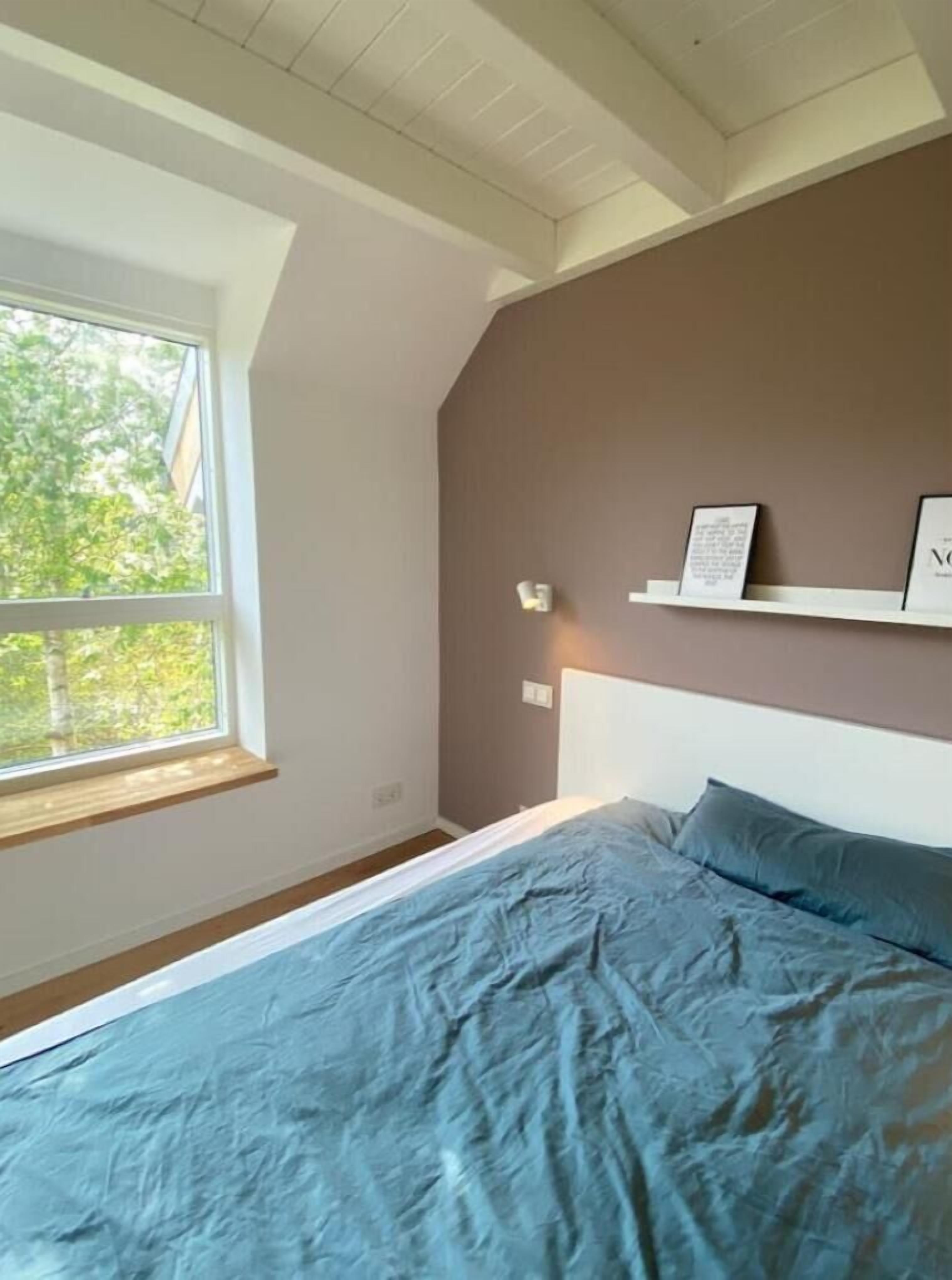 A bedroom with a sloped ceiling, a large window, a bed with blue bedding, and a shelf with framed items.