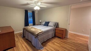 Een slaapkamer met een bed, houten vloer, plafondventilator, raam met gordijnen, nachtkastje en een dressoir.