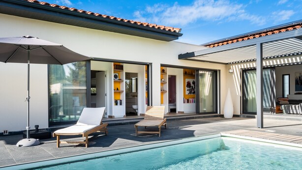 A modern outdoor pool area with two lounge chairs and a large umbrella, surrounded by a white building with large glass doors and a tiled roof.