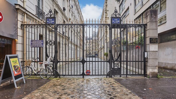 A gated entrance with a bicycle parked beside it, a signboard, and a cobblestone pathway leading to a street.