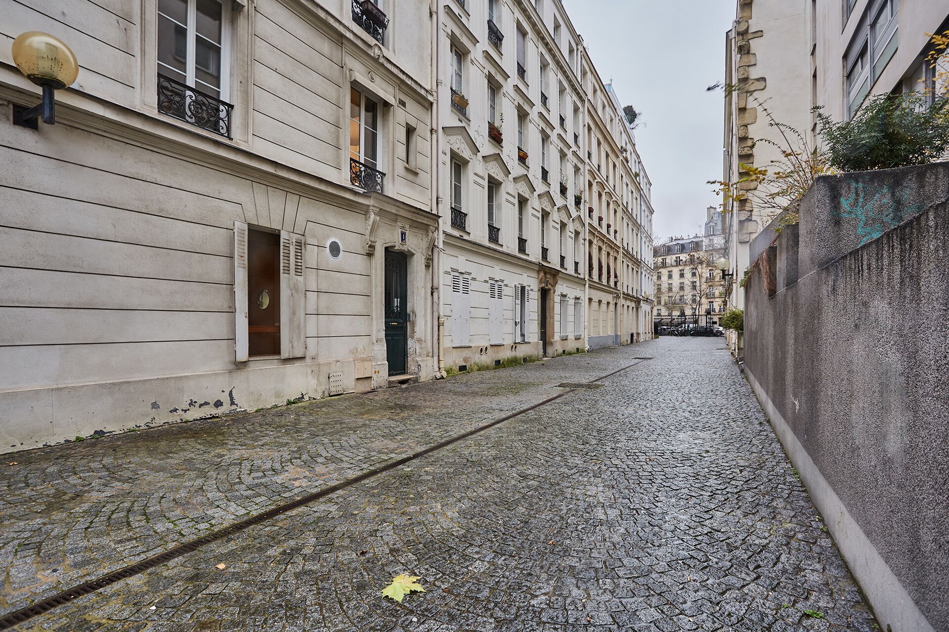 A narrow cobblestone street flanked by historic buildings with classic architecture, large windows, and decorative iron railings.