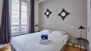A hotel room with a bed, two wall-mounted mirrors, a window with curtains, and a bedside table with a lamp.