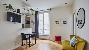A modern living room with a yellow armchair, a dining area with a table and chairs, a kitchen with a TV and plants, and a large window with curtains.