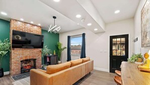 A modern living room with a brick fireplace, leather sofa, wooden dining table, and a large TV mounted on the wall. - Colorful Home with Deck • Near Acrisure Stadium & Downtown (Pittsburgh)