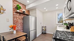 A modern kitchen with a brick accent wall, stainless steel appliances, and a dining area with a wooden table and benches. - Colorful Home with Deck • Near Acrisure Stadium & Downtown (Pittsburgh)