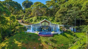 Une maison moderne dotée d’une grande façade en verre, entourée d’une végétation luxuriante et agrémentée d’une piscine.