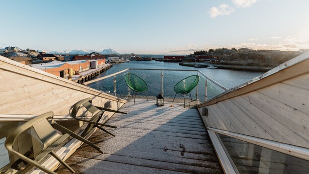 A deck with chairs overlooking a harbor, wooden railings, and a view of the water and distant buildings. - In the middle of Lofoten! Unique and spacious dockside apartment. (STAMSUND)